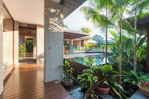 A modern outdoor pool area with a wooden deck, surrounded by tropical plants and a glass-enclosed swimming pool. - Luxury 5-Bed Beachfront Mirage Villa with Pool (Port Douglas)