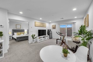 Una sala moderna con un sofá gris, una mesa de comedor y un televisor. Hay un dormitorio con una cama gris y un televisor. La habitación tiene paredes y techo blancos, además de plantas y obras de arte en las paredes.