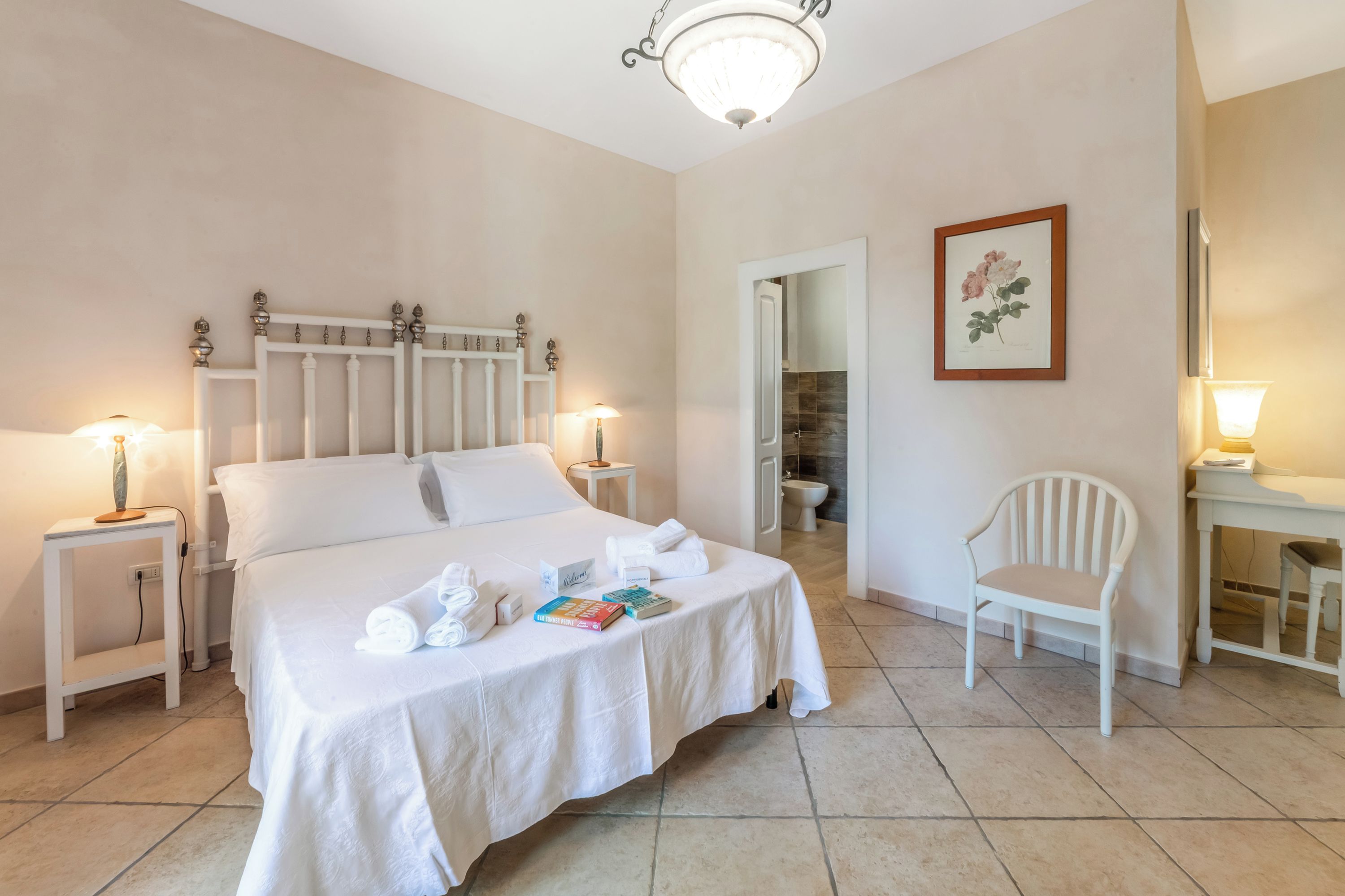 A hotel room with a large bed, bedside lamps, a chair, a desk, and a framed botanical print on the wall.