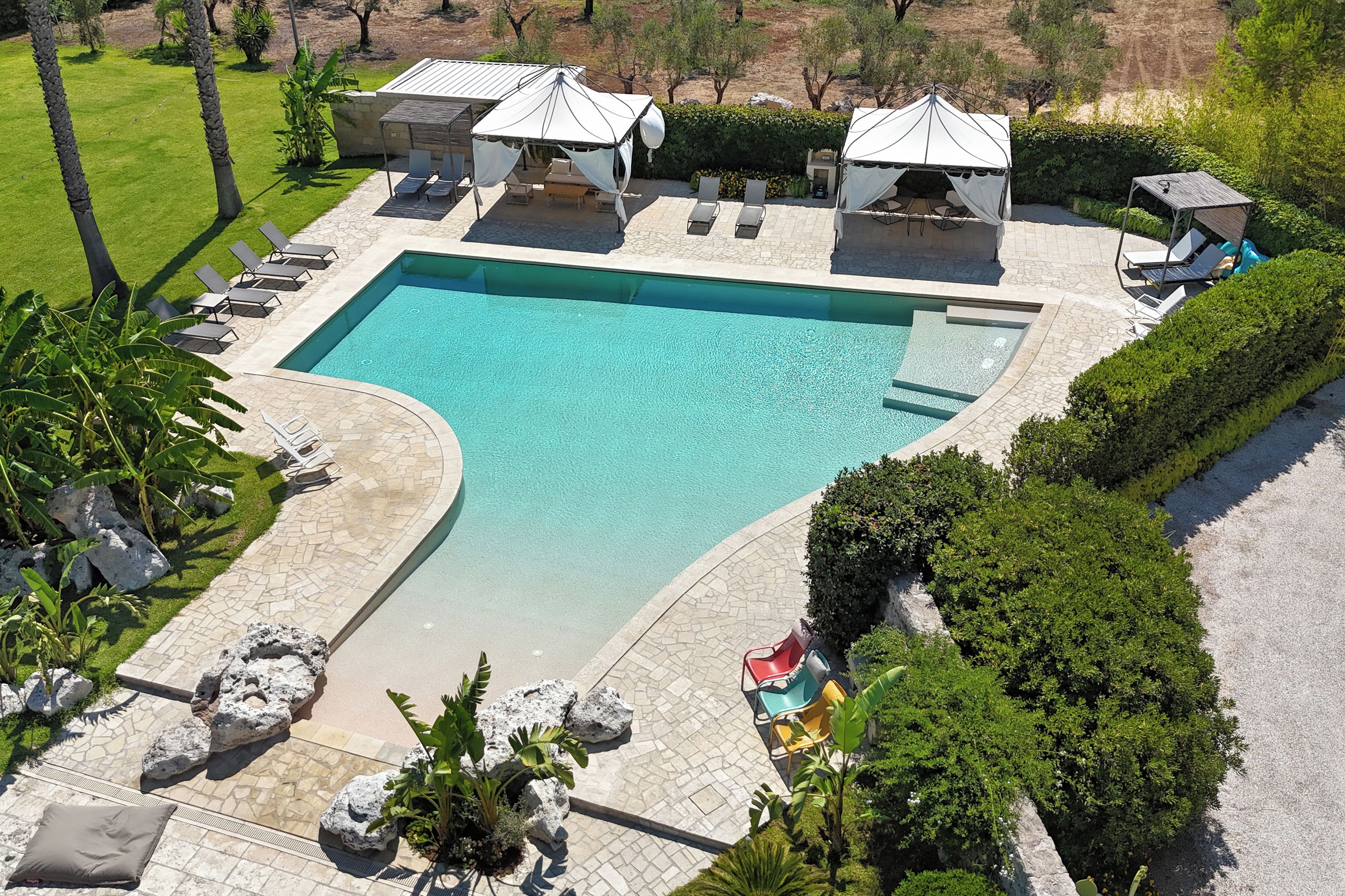 Una amplia piscina con sillones de descanso y sombrillas, rodeada de jardines ajardinados y un paisaje montañoso de fondo.