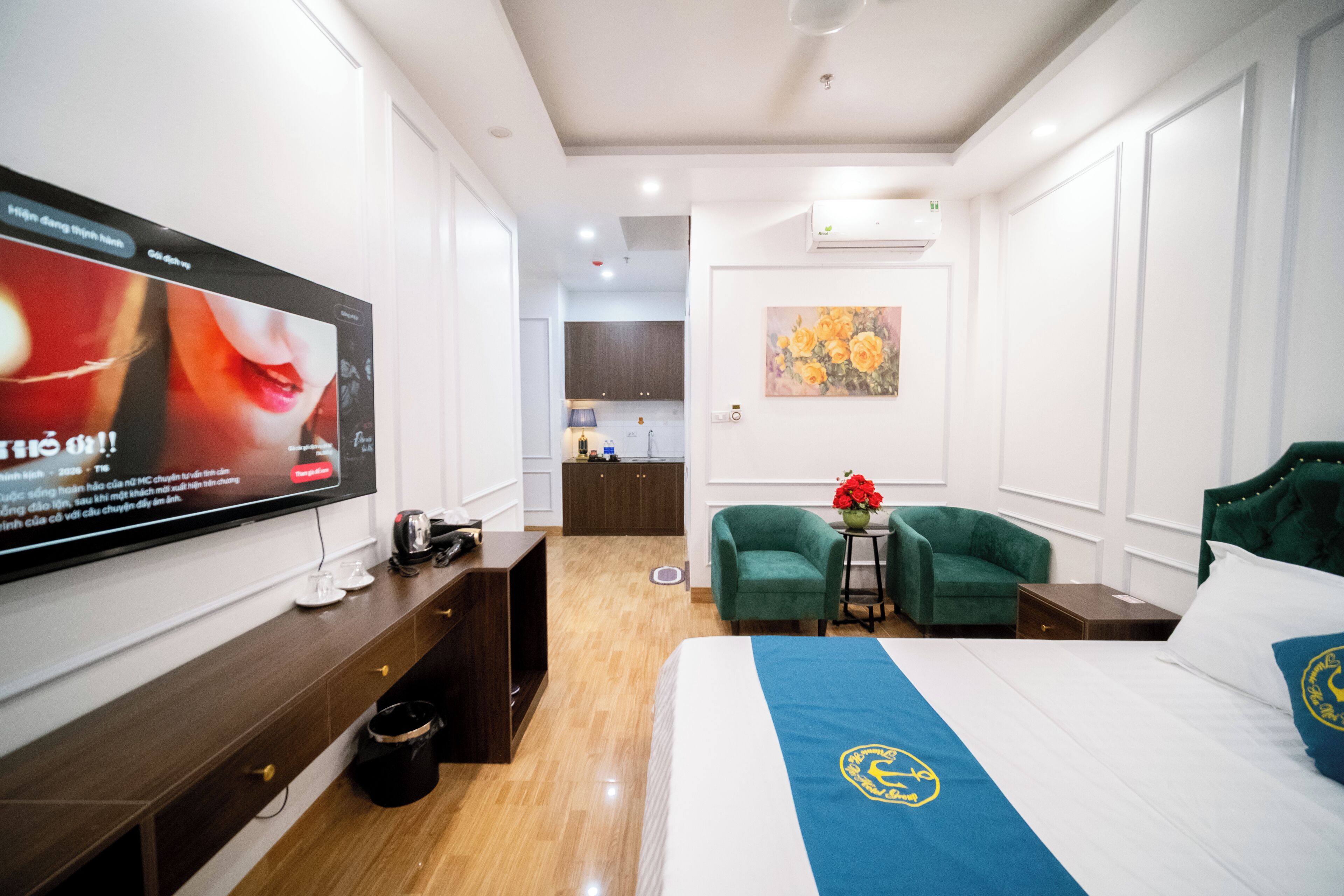 Habitació d'hotel amb un televisor de pantalla plana gran penjat a la paret, un llit amb edredó blau i blanc, un sofà verd i una taula petita amb un ram de flors.