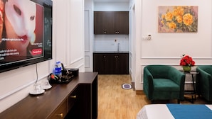 Een hotelkamer met een televisie, een bureau met een afstandsbediening, een bijzettafeltje met een bloemenvaas, een groene fauteuil en een schilderij aan de muur.