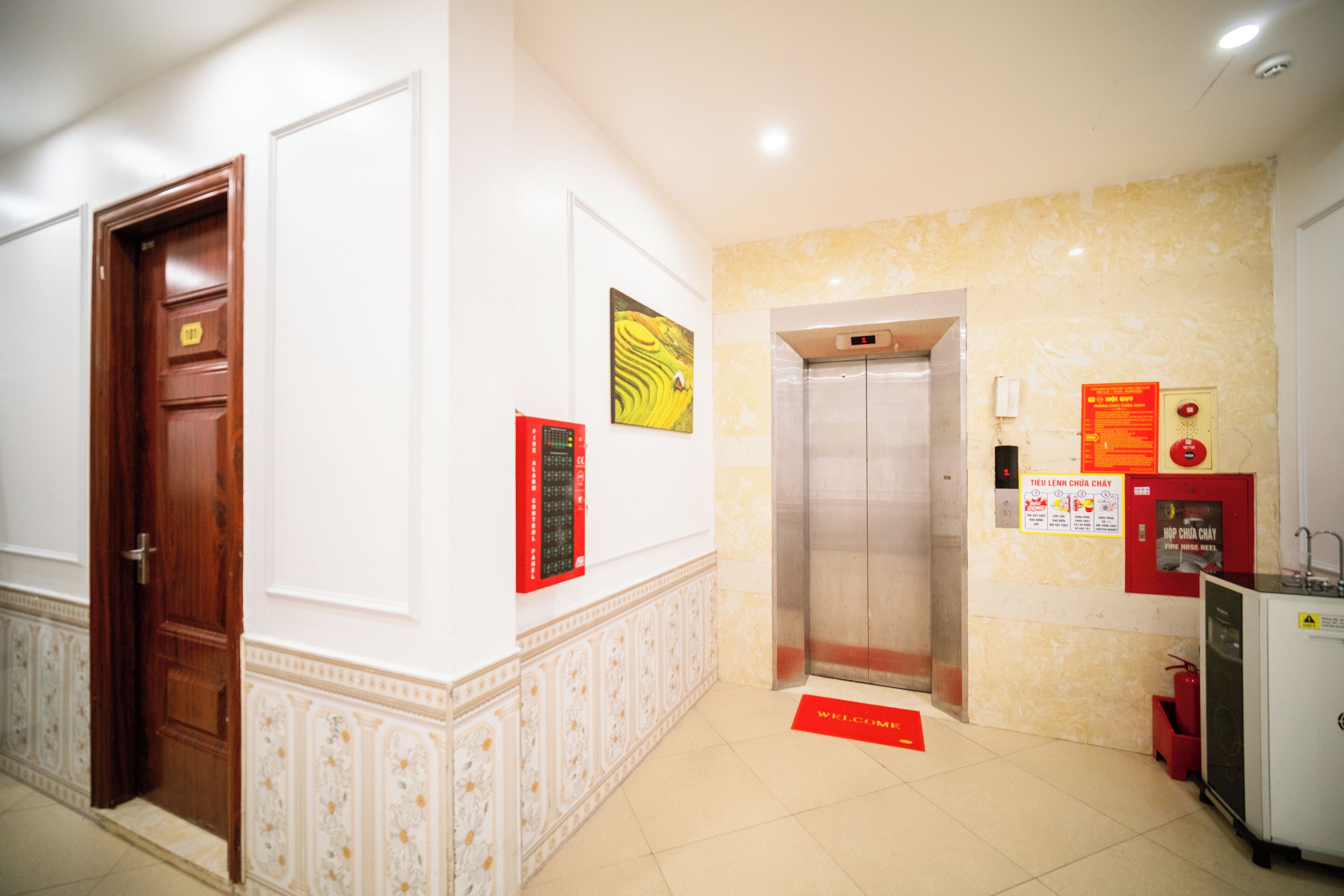 Un vestíbulo de hotel con una puerta de madera, un ascensor empotrado en la pared y una alfombra de bienvenida.