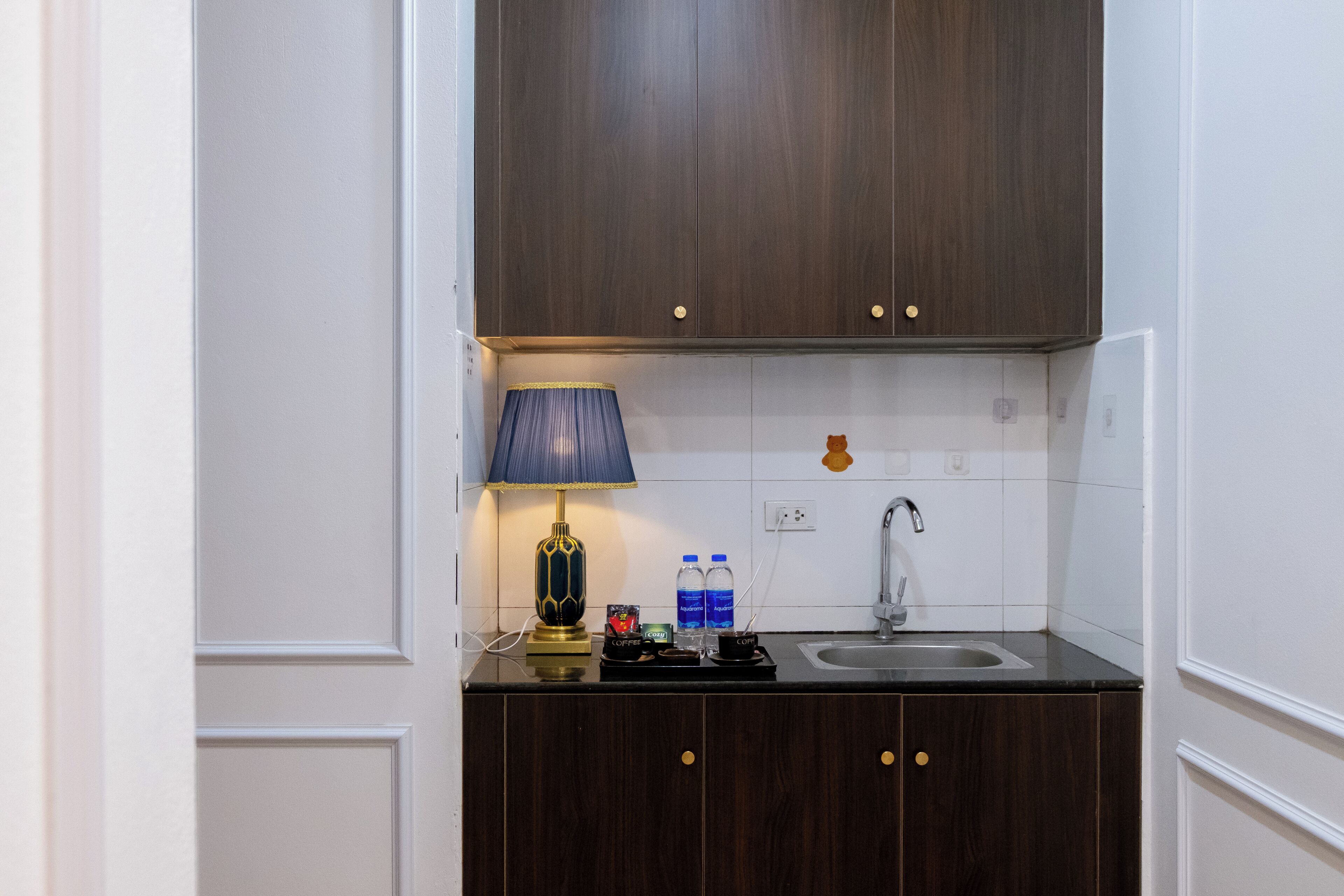 A compact kitchen with dark wood cabinets, a white tiled backsplash, a single faucet, and a small countertop with a lamp, two water bottles, and a coffee maker.