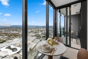 A modern dining area with a table set for two, overlooking a cityscape through large windows. - Luxury 1BR with Stunning Ocean Views (Broadbeach)