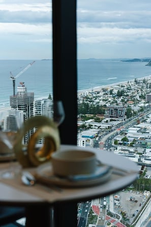 A table with a cup and saucer, a plate, and a spoon, with a view of a coastal cityscape and the ocean through a window. - Luxury 1BR with Stunning Ocean Views (Broadbeach)