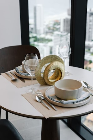 A dining table set with plates, cutlery, and a decorative bowl, with a cityscape view through a window. - Luxury 1BR with Stunning Ocean Views (Broadbeach)