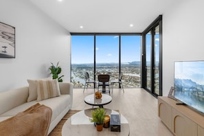 A modern living room with a sofa, coffee table, and chairs, overlooking a cityscape through large windows. - Luxury 1BR with Stunning Ocean Views (Broadbeach)