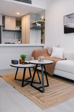 A modern living room with a sofa, a coffee table, and a small side table with a plant and a box. - Luxury 1BR with Stunning Ocean Views (Broadbeach)
