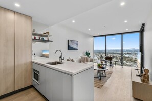 A modern kitchen with a large island, a dining area with a view, and a living space with a large window. - Luxury 1BR with Stunning Ocean Views (Broadbeach)