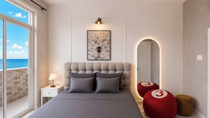 A bedroom with a large bed, a nightstand, a lamp, a framed picture, a mirror, and a view of the ocean. - MOC AN COLLECTION-Blue Sea Beachfront VT (Vung Tau)