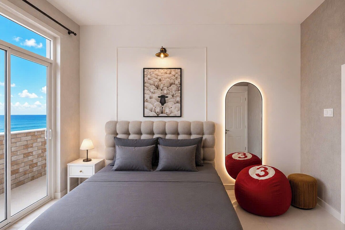 A bedroom with a large bed, a nightstand, a lamp, a framed picture, a mirror, and a view of the ocean.
