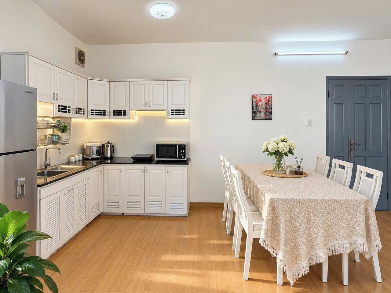 Cocina compacta con gabinetes blancos, microondas y lavamanos. Hay una mesa de comedor con sillas y un florero con flores.