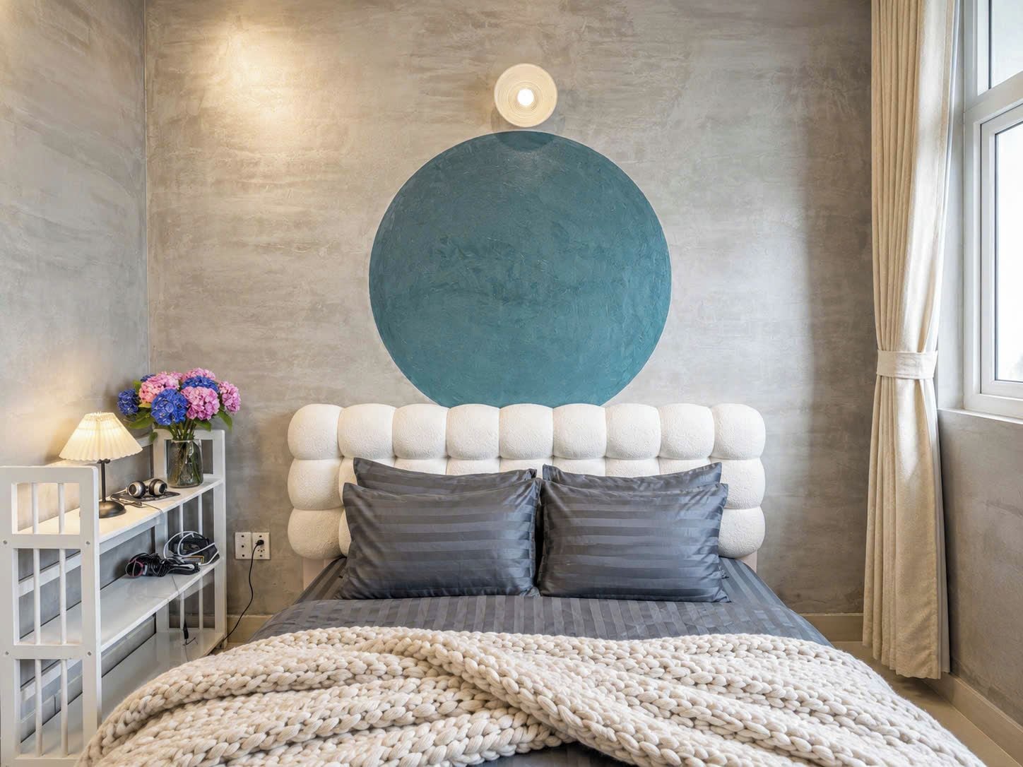 Quarto com cabeceira estofada, uma grande decoração circular na parede, mesa de cabeceira com abajur e cama com manta de malha.