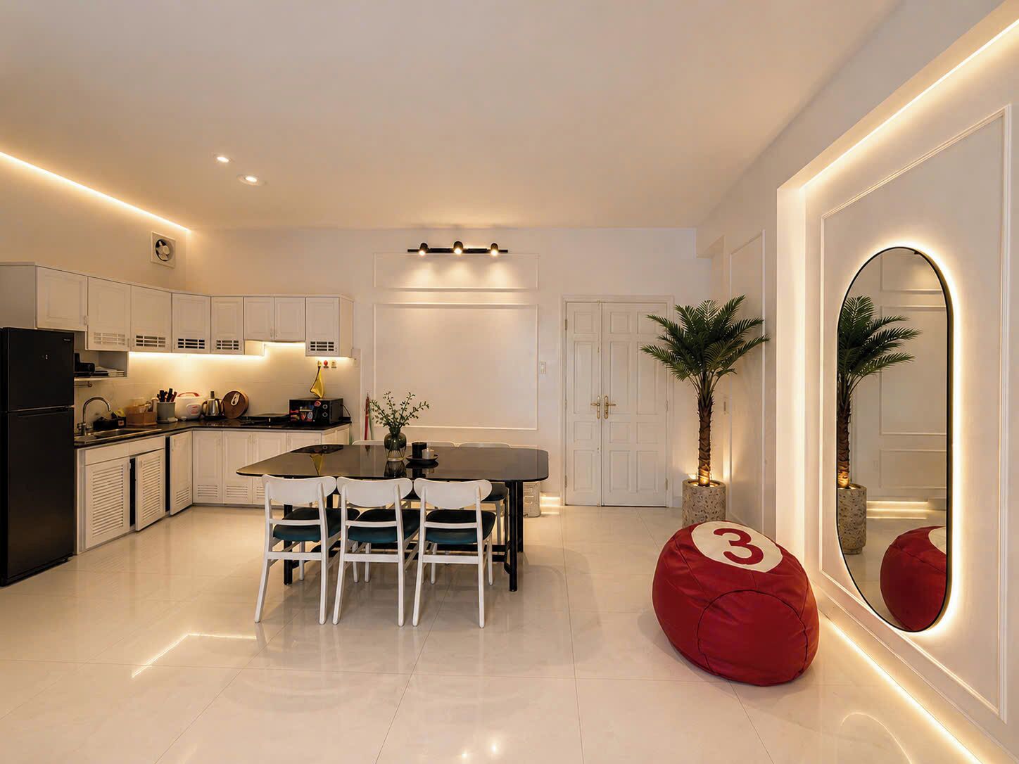 A modern kitchen with white cabinetry, a dining table with chairs, a red bean bag, and a large mirror with a number '3' on it.