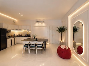 A modern kitchen with white cabinetry, a dining table with chairs, a red bean bag, and a large mirror with a number '3' on it. - MOC AN COLLECTION-Blue Sea Beachfront VT (Vung Tau)