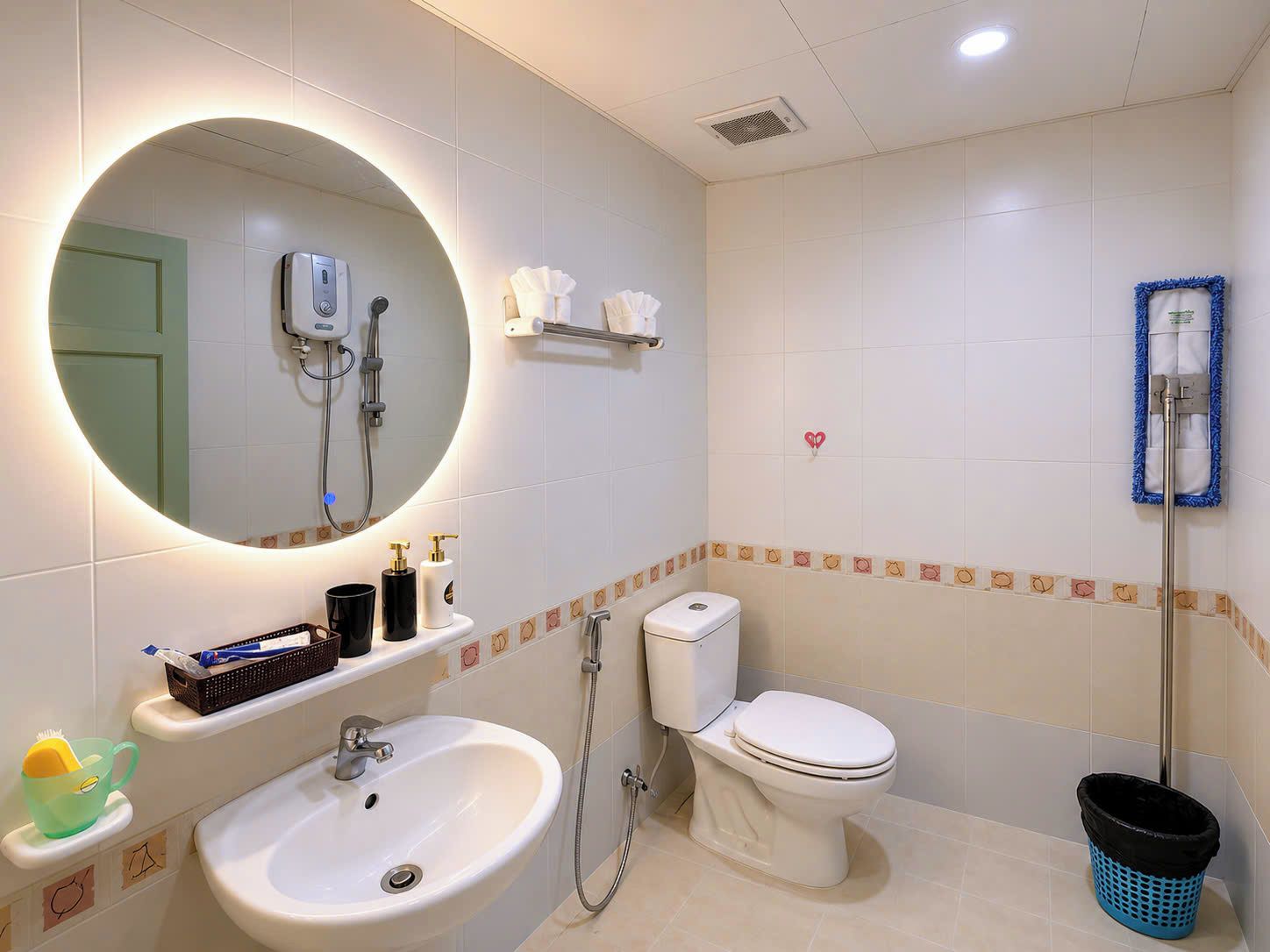 Baño con lavamanos, inodoro y ducha. Se observa un espejo con luz, un perchero y un bote de basura.