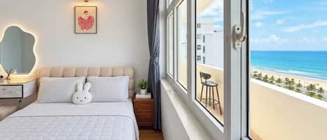 A bedroom with a large window, a bed with a white quilt, a bedside table with a lamp, and a view of the beach.