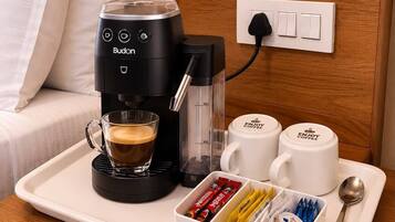 Una cafetera, una taza de café, dos tazas, una bandeja con cápsulas de café y una cuchara sobre la mesita de noche de una habitación de hotel.