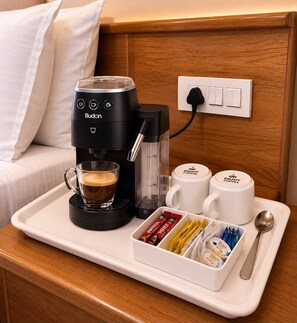 Uma cafeteira, um copo de café, duas canecas, um suporte com cápsulas de café e uma colher em uma mesa de cabeceira de quarto de hotel.