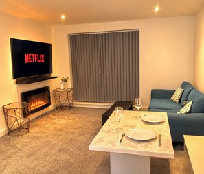 Ein Wohnzimmer mit Kamin, einem flachbildfernseher, der darüber angebracht ist, einem Sofa, einem Couchtisch mit Tellern und Besteck sowie einem Fenster mit Jalousien.