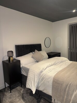 A modern bedroom with a bed, nightstands, a lamp, and a window with blinds. - Cosy Modern 3 Bedroom Apartment with Secure Private Carpark (Leeds)
