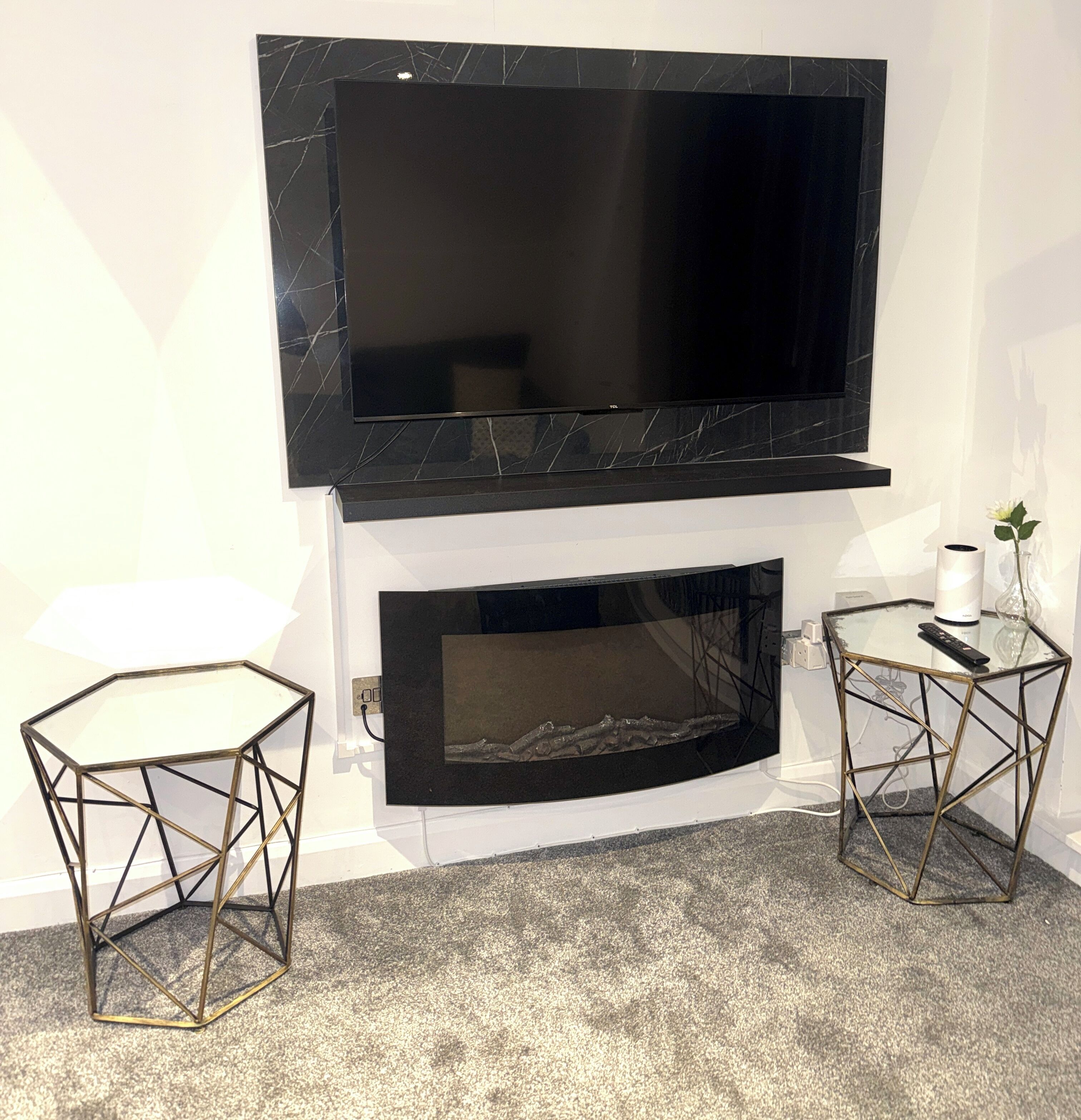 A modern living room with a flat-screen TV mounted above a fireplace, two glass tables with metal frames, and a carpeted floor.