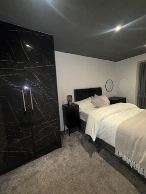 A bedroom with a large wardrobe, a bed with white bedding, a nightstand with a lamp, and a circular wall decoration. - Cosy Modern 3 Bedroom Apartment with Secure Private Carpark (Leeds)