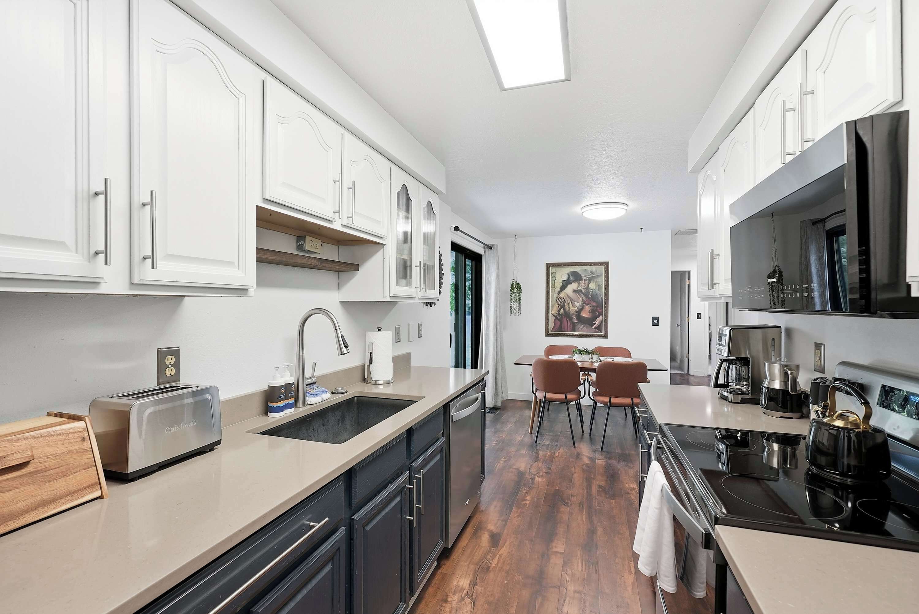 Una cocina compacta con muebles blancos, horno empotrado, cafetera y un comedor con mesa y sillas.