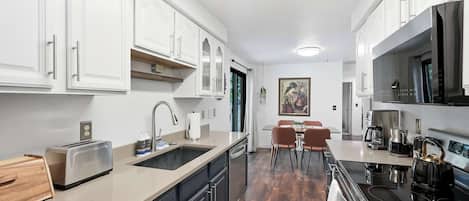 Una cocina compacta con muebles blancos, horno empotrado, cafetera y un comedor con mesa y sillas.