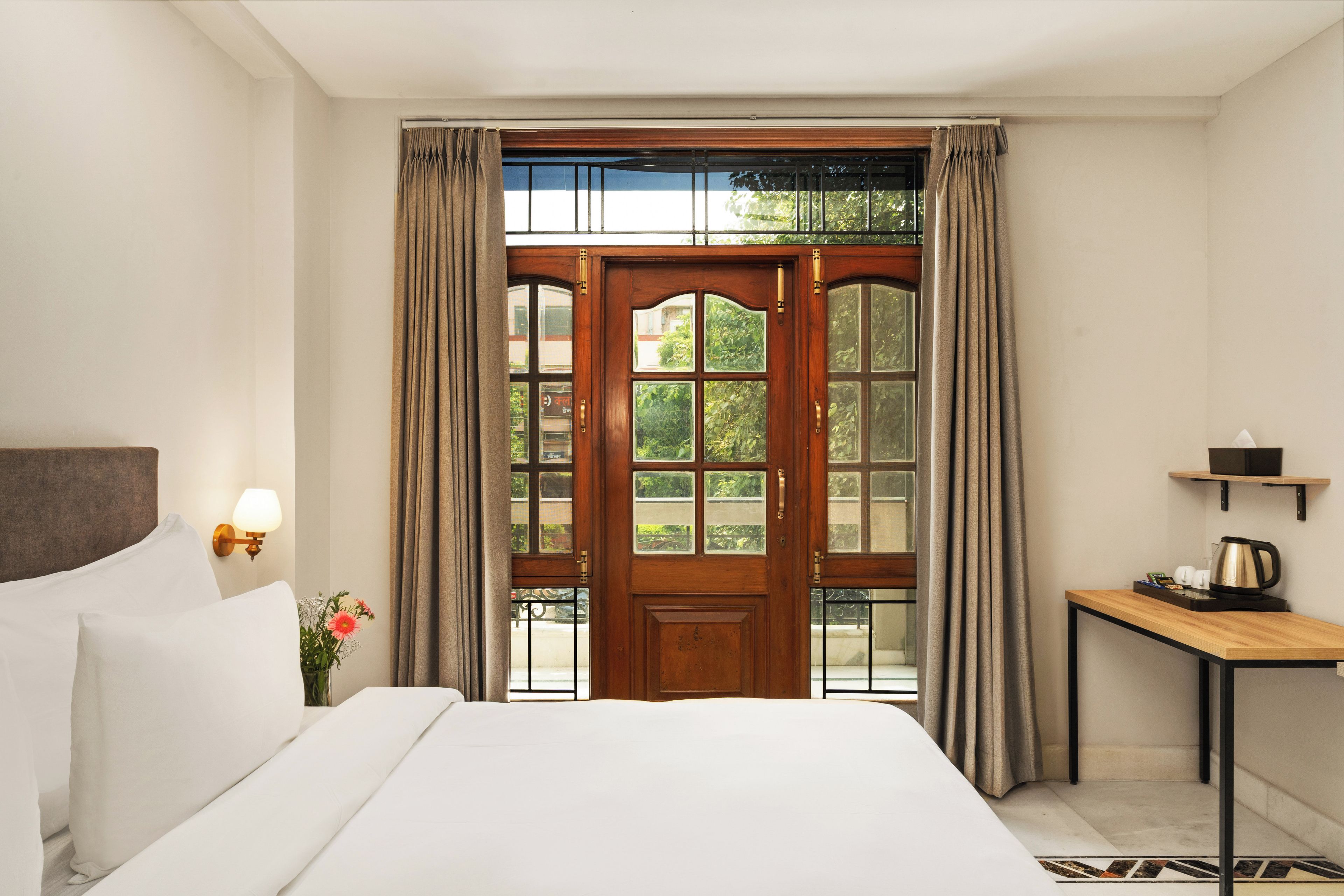 Una camera d'albergo con un letto grande, una porta in legno con pannelli di vetro, un tavolino con un bollitore e un comodino con una lampada.