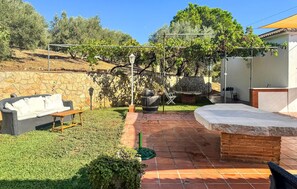 Eine Terrasse mit Steinmauer, einem Korbssessel, einem Holztisch und einem Grill.
