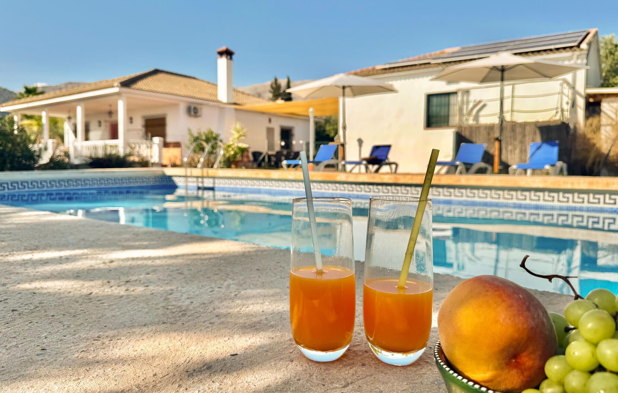 A poolside setting with two glasses of amber liquid, a fruit bowl with grapes and a peach, and a building with a tiled roof in the background.