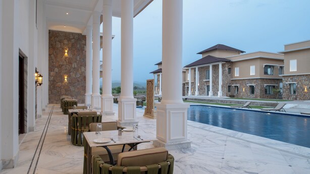 A covered outdoor area with marble flooring, dining tables, and chairs, overlooking a swimming pool and a large building with stone walls. - Hari Bagh Jawai (Bali)
