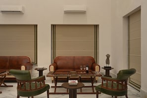 A room with a brown leather sofa, two green upholstered chairs, a small round table with a wine bottle and glasses, and a decorative sculpture on a pedestal.