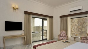 A hotel room with a bed, a television, a desk, a chair, a small table with a fruit bowl, and a stone wall. - Hari Bagh Jawai (Bali)