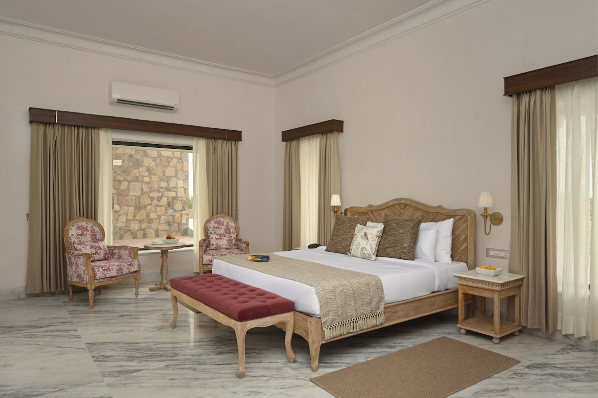 Een hotelkamer met een groot bed, twee stoelen, een kleine tafel, een stenen muur en uitzicht op de omgeving.