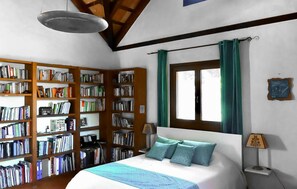Ein Schlafzimmer mit einem großen Bett, einem Bücherregal, einem Fenster mit Vorhängen und einer Lampe.