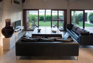 Ein modernes Wohnzimmer mit Sofa, Couchtisch und Fernseher. Große Fenster bieten Blick auf einen Garten.