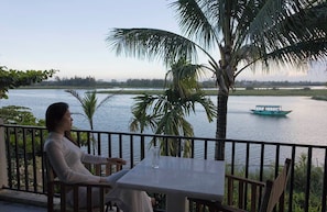 Une femme est assise à une table, devant laquelle s’étend une rivière et sur laquelle on aperçoit un bateau.