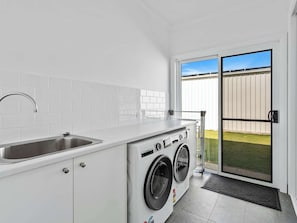 A compact laundry room with a sink, washing machine, and a sliding glass door leading to a backyard. - The Hamptons Retreat - Spring Offer (Yarrawonga)