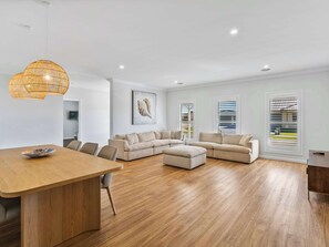 A modern living room with a wooden dining table, a sectional sofa, and a large window with blinds. - The Hamptons Retreat - Spring Offer (Yarrawonga)