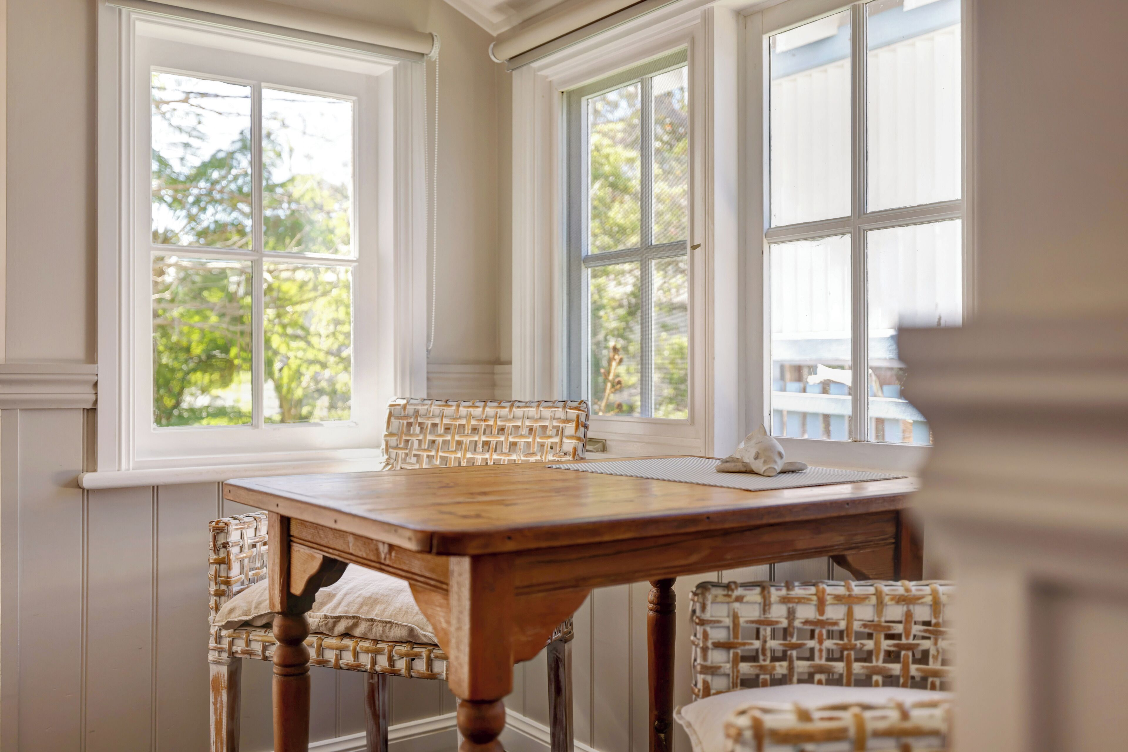 A dining area with a wooden table and wicker chairs, large windows with a view of greenery, and a white ceiling.