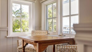 Et spiseområde med et træbord og fletstole, store vinduer med udsigt over grøn vegetation, og et hvidt loft.