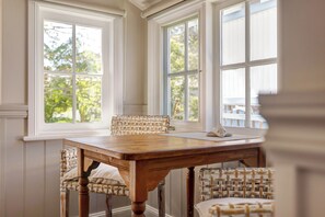Comedor con mesa de madera y sillas de mimbre, ventanas amplias con vista a vegetación y techo blanco.