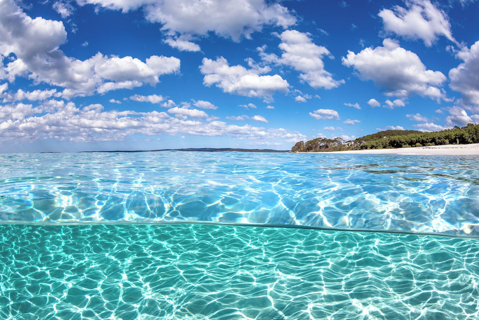 A clear blue sea with a sandy beach and a forested hill in the background.