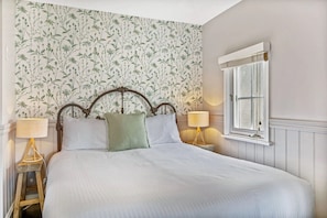 Ein Schlafzimmer mit floralem Wandpapier, einem klassischen Bettgestell, zwei Nachttischlampen und einem Fenster mit Fensterläden.
