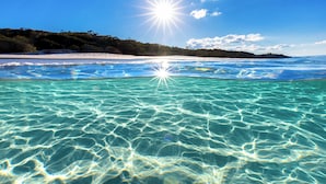 En strand med krystalklart turkis vand, en hvid sandstrand og en skovklædt bakke i baggrunden.