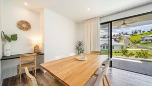Uma sala de jantar moderna com mesa de madeira, cadeiras e vista para um bairro residencial através de grandes janelas.