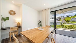 A modern dining area with a wooden table, chairs, and a view of a residential neighborhood through large windows. - Habitat - Lush Lennox Head Apartment (Lennox Head)